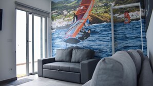 A living room with a large mural of a windsurfer on water, a grey sofa, and a sliding glass door leading to an outdoor area. - Hostel AzorAzul (Ponta Delgada)