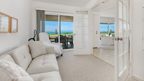 Un salon moderne avec un canapé blanc, un coin repas comprenant une table et des chaises, et une grande porte-fenêtre coulissante donnant sur un balcon avec vue sur la mer.
