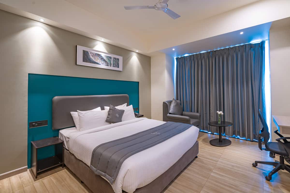 Quarto de hotel moderno com uma cama grande, uma parede de destaque cor turquesa, um ventilador de teto, uma mesa com cadeira e uma janela com cortinas.