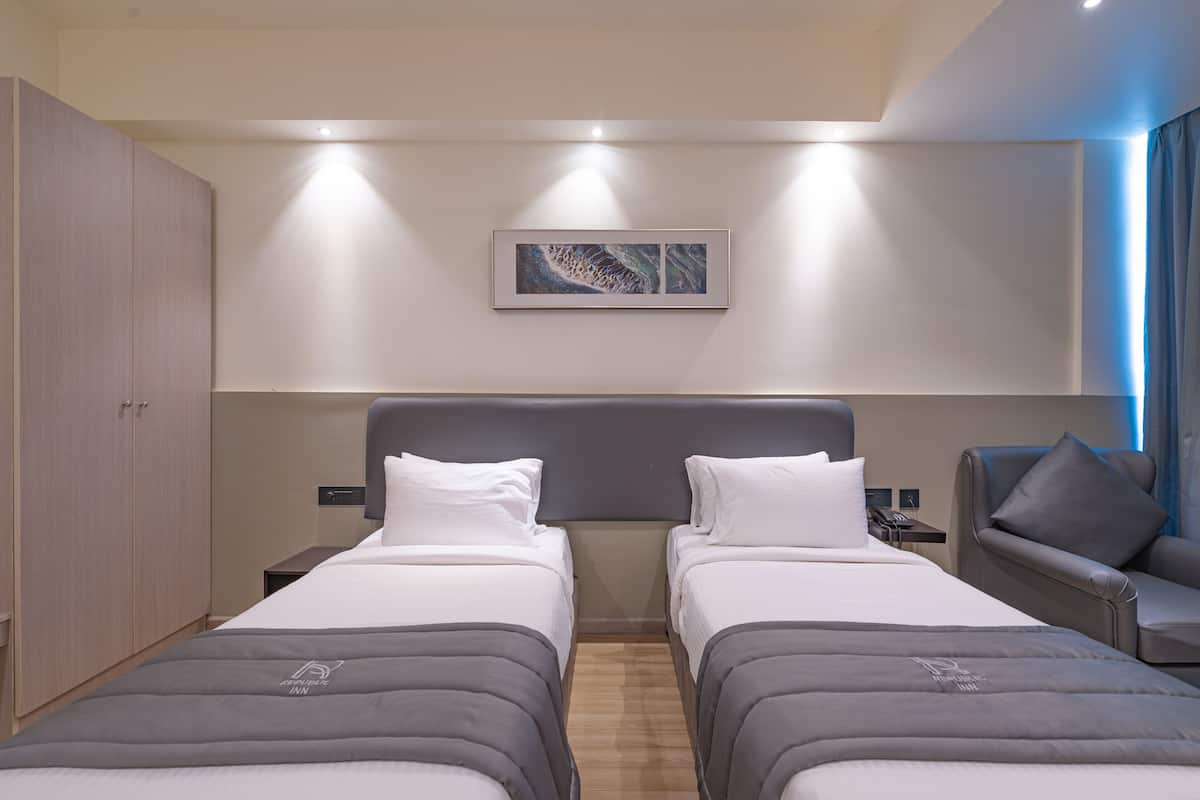 Quarto de hotel com duas camas, cabeceira cinzenta, mesa de cabeceira, escrivaninha e uma cadeira.