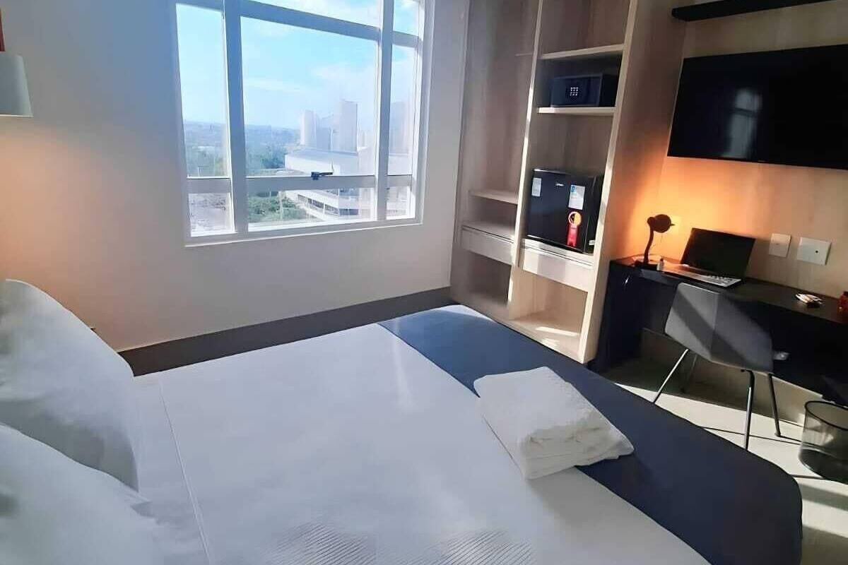 A modern hotel room with a large bed, a desk with a chair, a TV, and a view of the city through a large window.