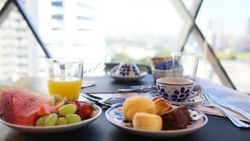 Una mesa de desayuno con sandía, uvas, fresas, un vaso de jugo de naranja, un plato con croissants y un postre de chocolate, y una taza de café.