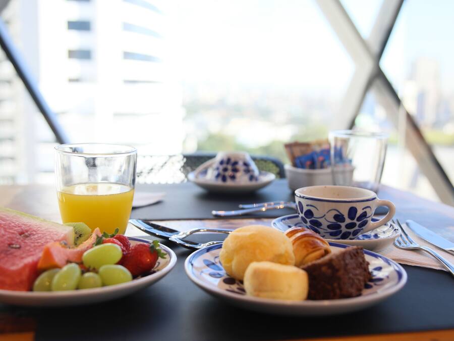 Una mesa de desayuno con sandía, uvas, fresas, un vaso de jugo de naranja, un plato con croissants y un postre de chocolate, y una taza de café.