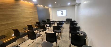 A conference room with rows of chairs and tables, a wooden accent wall, and a projector screen displaying a logo.