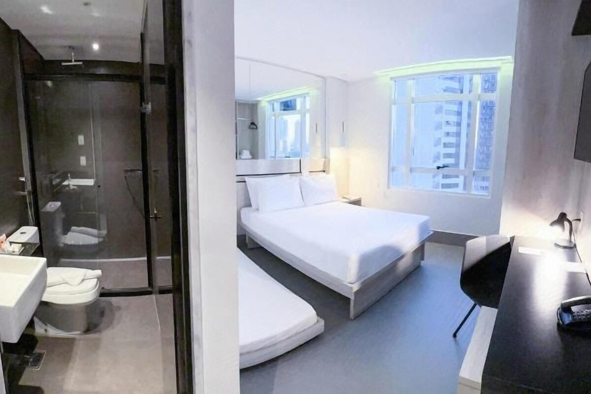 Una habitación de hotel moderna con cabina de ducha de vidrio, cama con ropa blanca, escritorio con lámpara y vista de la ciudad a través de grandes ventanales.