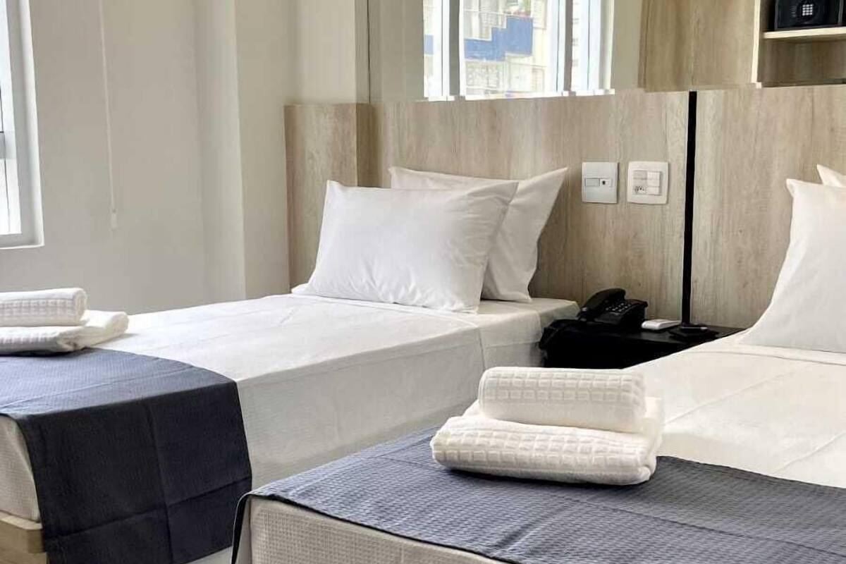 A hotel room with two beds, a large mirror, a wooden headboard, a bedside table with a phone, and a window with a view of buildings.
