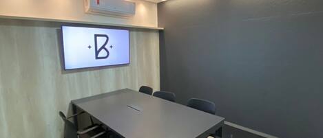 Un salón de conferencias con una mesa larga, sillas, una pantalla montada que muestra un logotipo y un equipo de aire acondicionado.