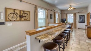 A kitchen with a long counter, bar stools, ceiling fans, and a window with curtains. - Aces & Eights - Downtown Sturgis with garage! (Sturgis)