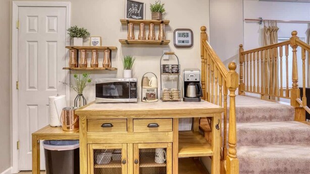 A kitchen with a wooden table, microwave, coffee maker, and shelves with decorative items. - Aces & Eights - Downtown Sturgis with garage! (Sturgis)