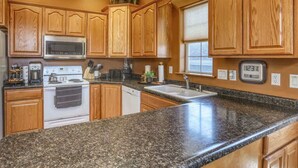 A kitchen with wooden cabinets, granite countertops, a microwave, stove, and a clock. - Aces & Eights - Downtown Sturgis with garage! (Sturgis)