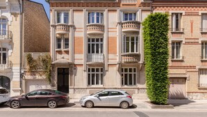 Un edificio con un balcón grande, un seto verde alto y dos coches estacionados.
