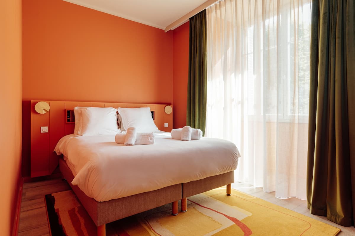Ein Hotelzimmer mit einem großen Bett, einem hölzernen Kopfteil, einem Nachttisch und einem Fenster mit transparenten Vorhängen.