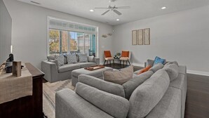 Ein modernes Wohnzimmer mit einem grauen Sofa, einem Couchtisch und einem Schreibtisch mit Lampe. Ein Deckenventilator und ein Fenster mit Blick auf Bäume sind vorhanden.