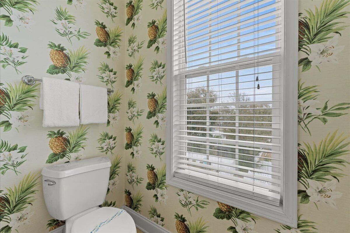 Ein Badezimmer mit Toilette, einem Fenster mit Jalousien und einer Wand mit Ananas- und Blumenmuster-Tapete.