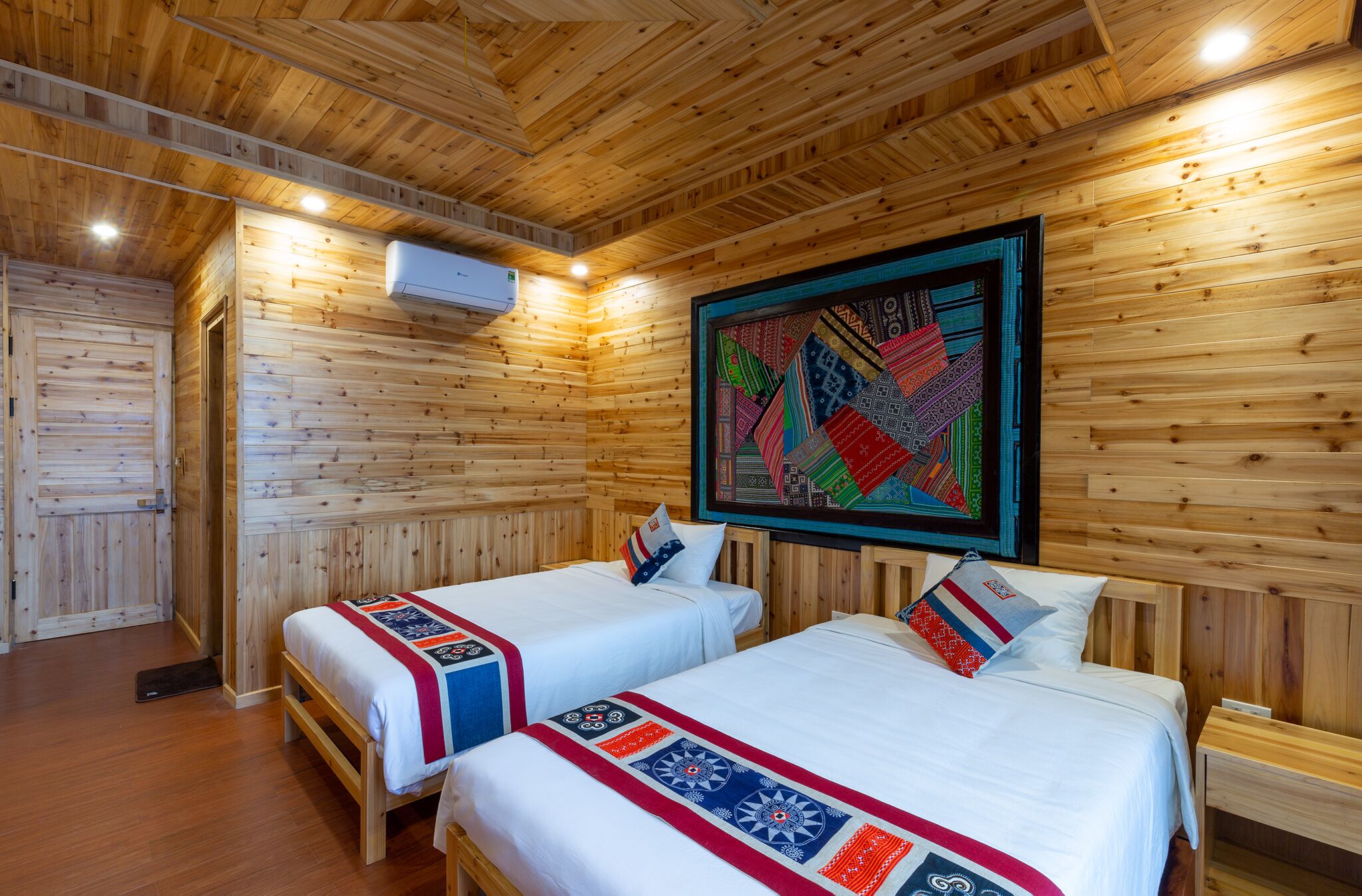 A wooden cabin room with two beds, a large colorful tapestry, and an air conditioning unit.