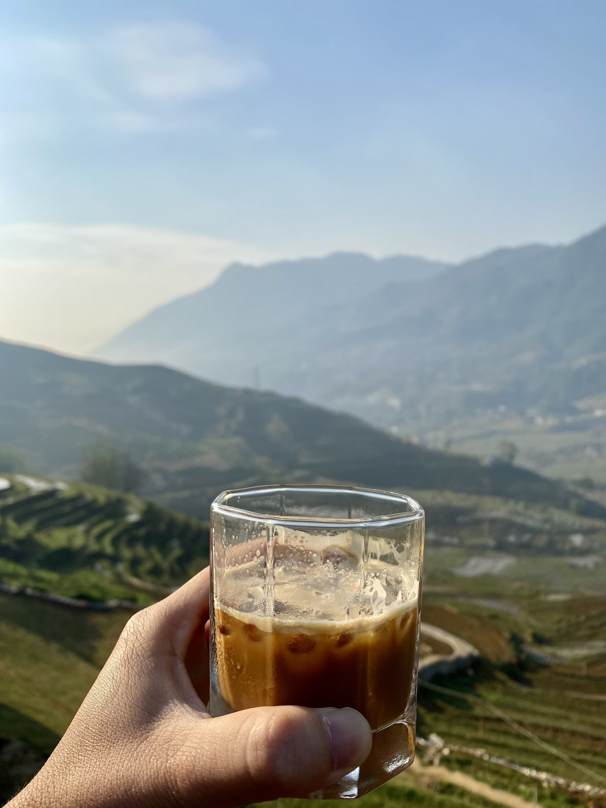 Una mano che tiene un bicchiere di caffè freddo, con un paesaggio montuoso sullo sfondo.
