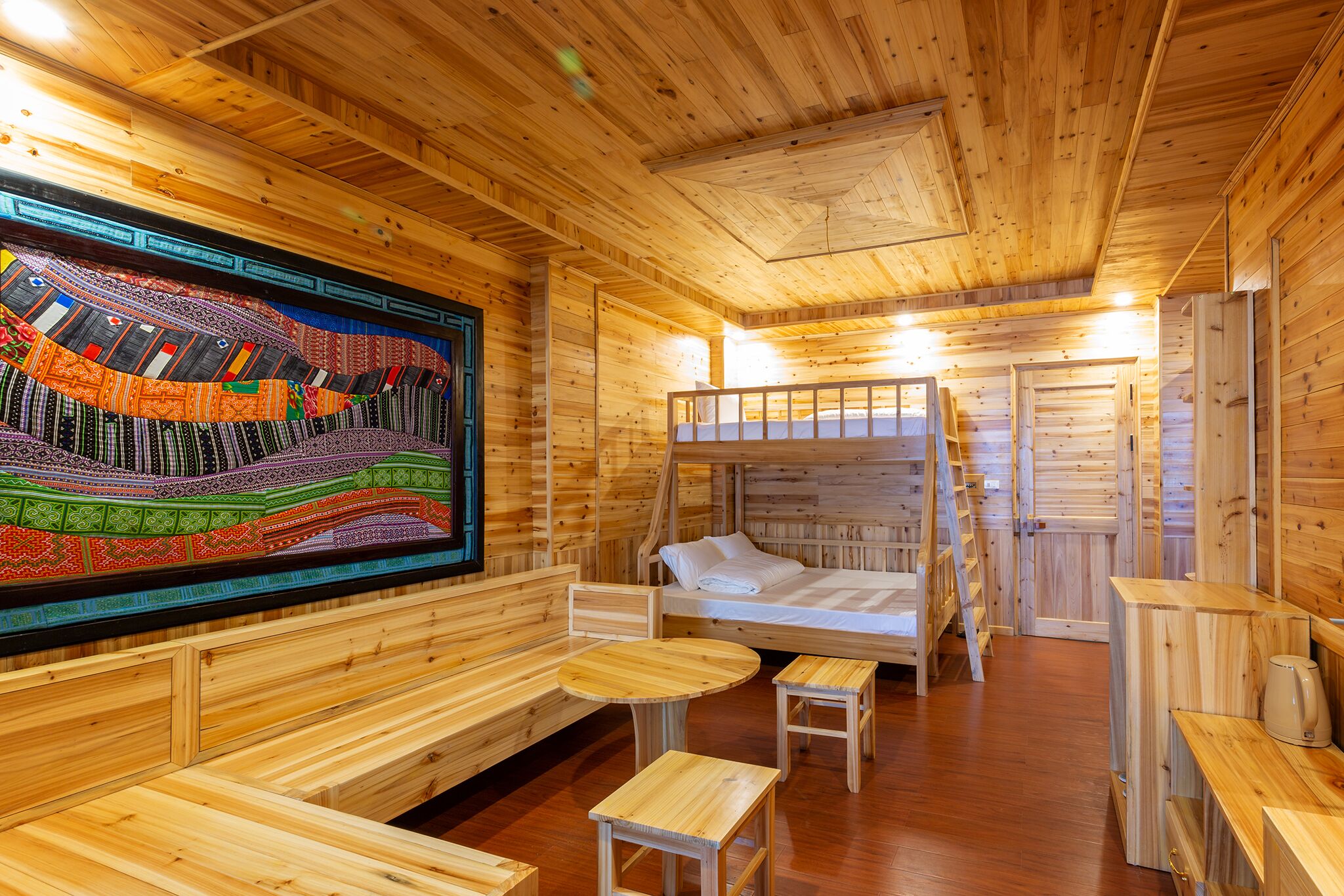 Uma cabana de madeira com uma cama beliche, um banco, uma mesa pequena e uma tapeçaria colorida.