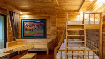 Interior de una cabaña de madera con literas, una mesa de comedor, bancos y un tapiz colorido en la pared.