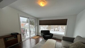 Un salon comprenant un canapé, une table basse, un fauteuil, un meuble pour la télévision et une grande fenêtre avec des stores.