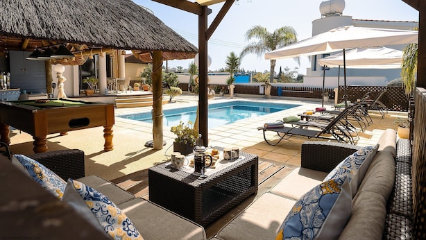 Un espace aménagé au bord de la piscine, comprenant une piscine, des chaises longues, une table avec des boissons et une structure recouverte d’un toit de chaume.