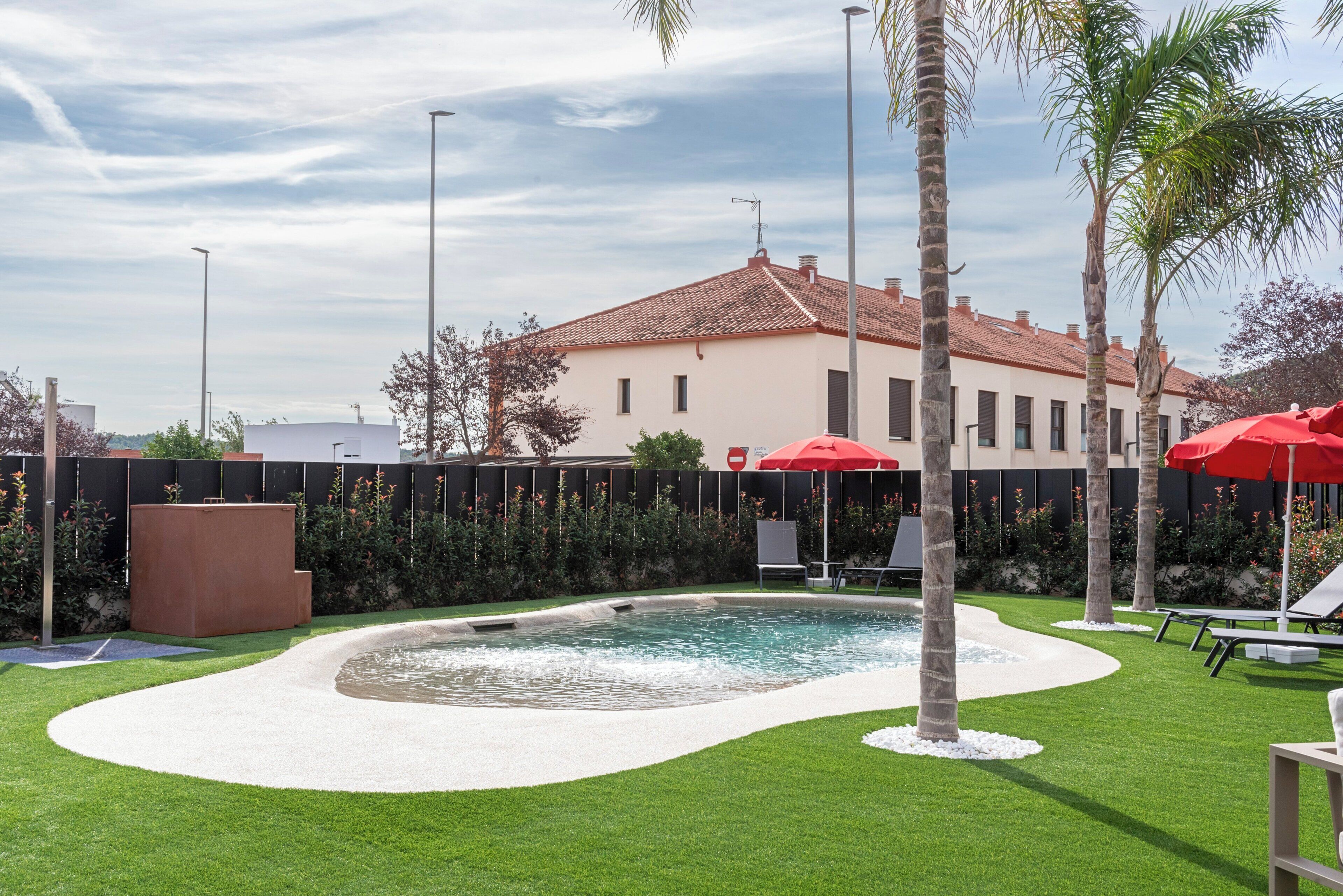 Un espace piscine doté d’une terrasse blanche, de palmiers et de parasols rouges.
