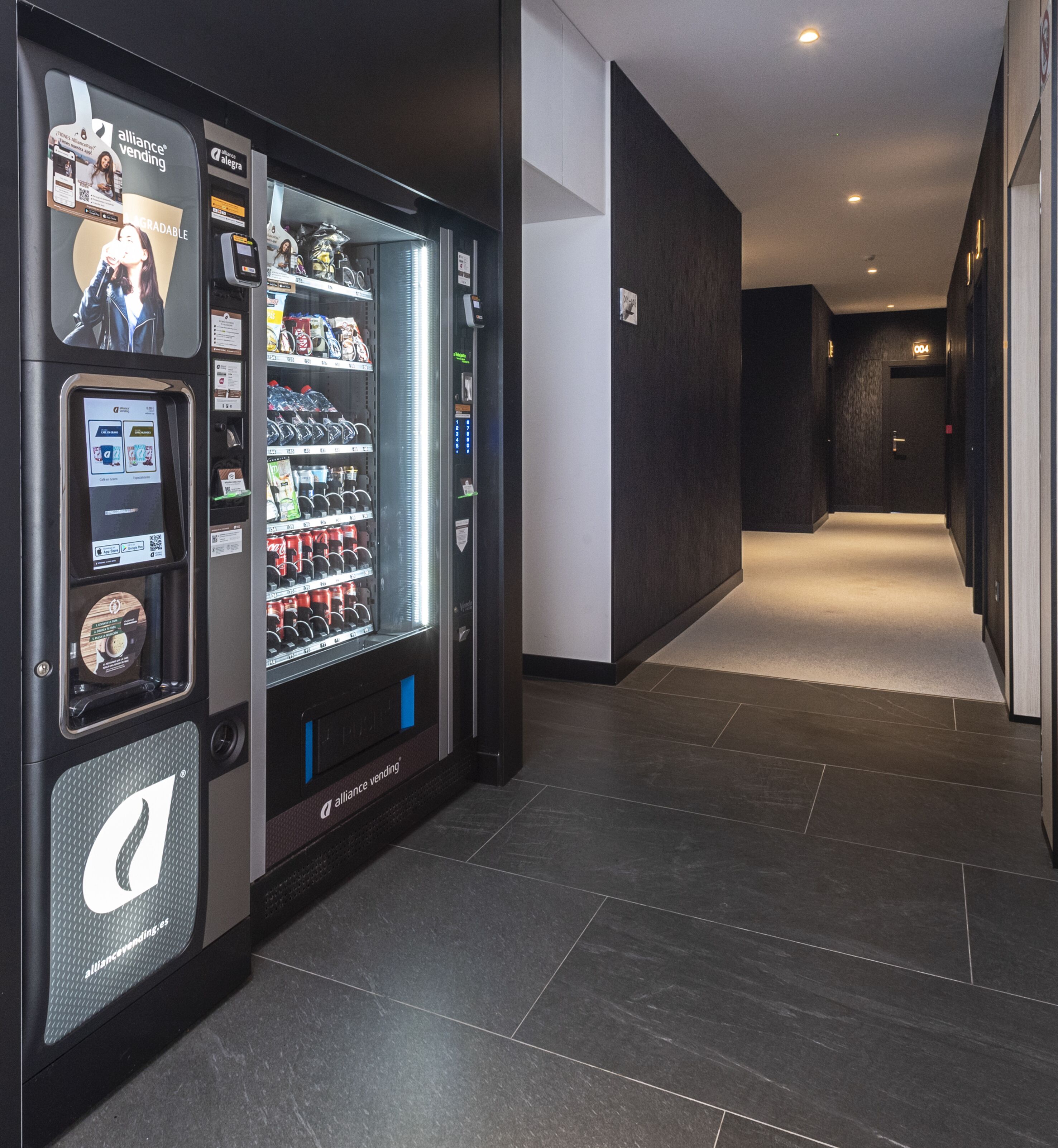 Una máquina expendedora con pantalla digital y un código QR, ubicada en un pasillo con piso de baldosas oscuras y diseño moderno.