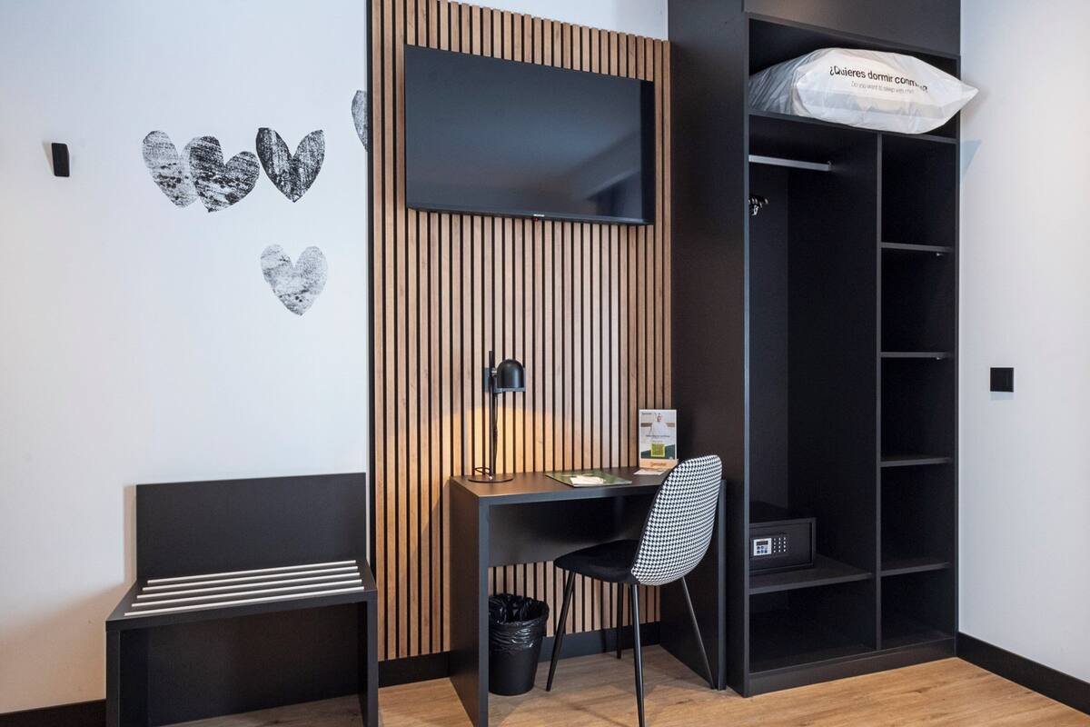 Una habitación moderna con un cabecero de listones de madera, un escritorio con silla, un estante negro y un cubo de basura negro.