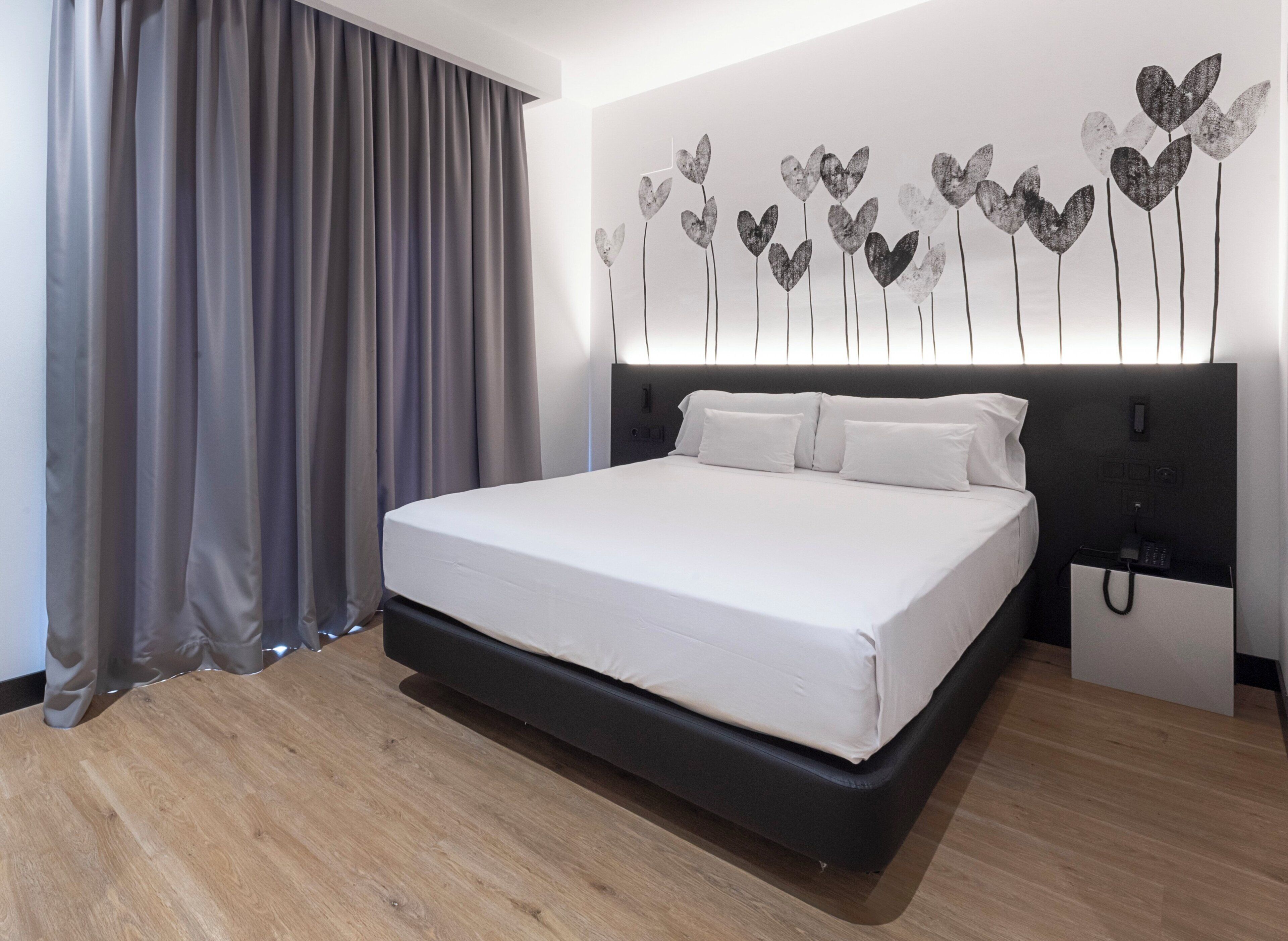 A modern hotel room with a large bed, grey curtains, and a wall-mounted light fixture with heart-shaped decorations.