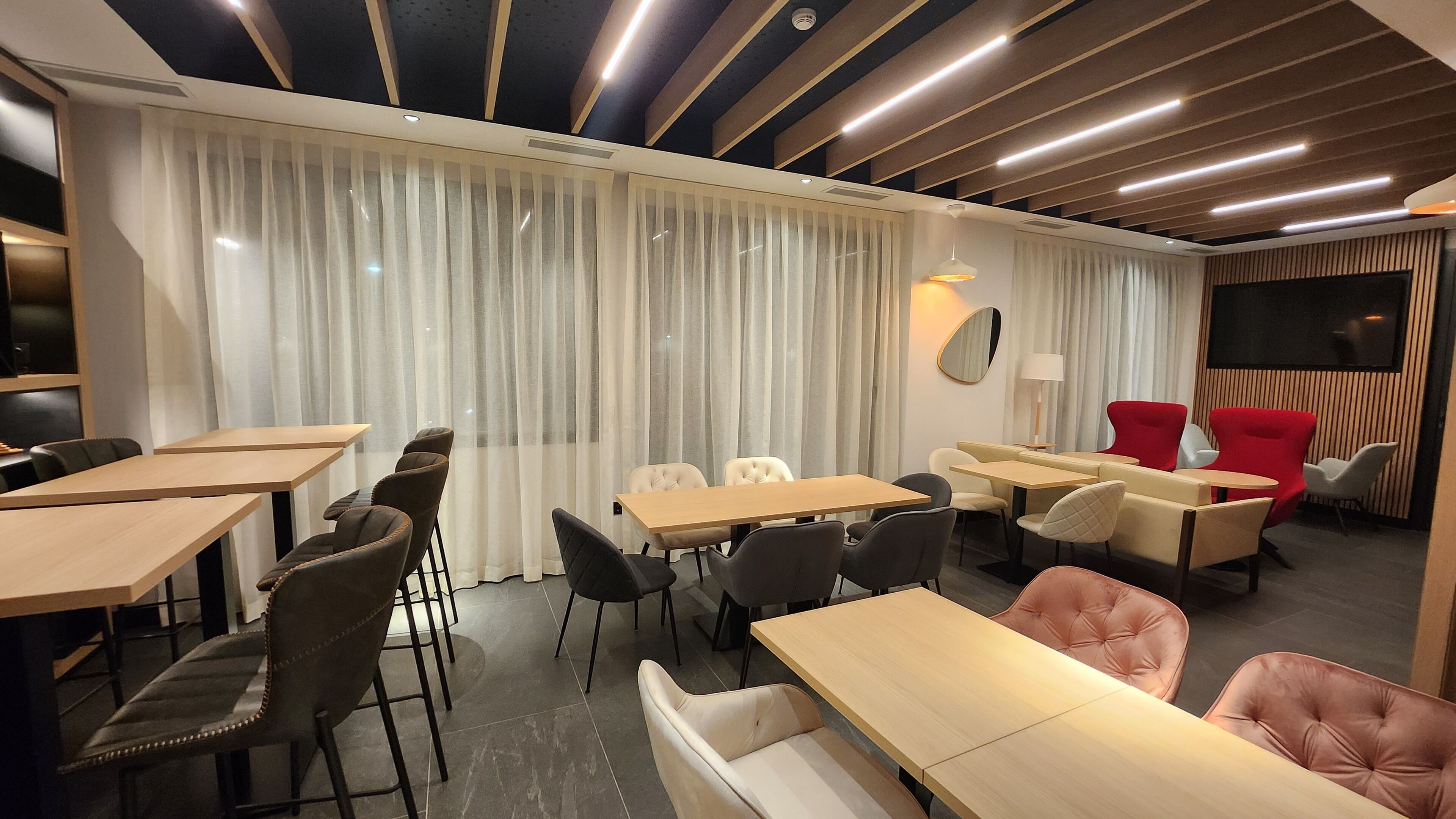 A modern dining area with wooden tables and chairs, a large window with curtains, and a wall-mounted TV.