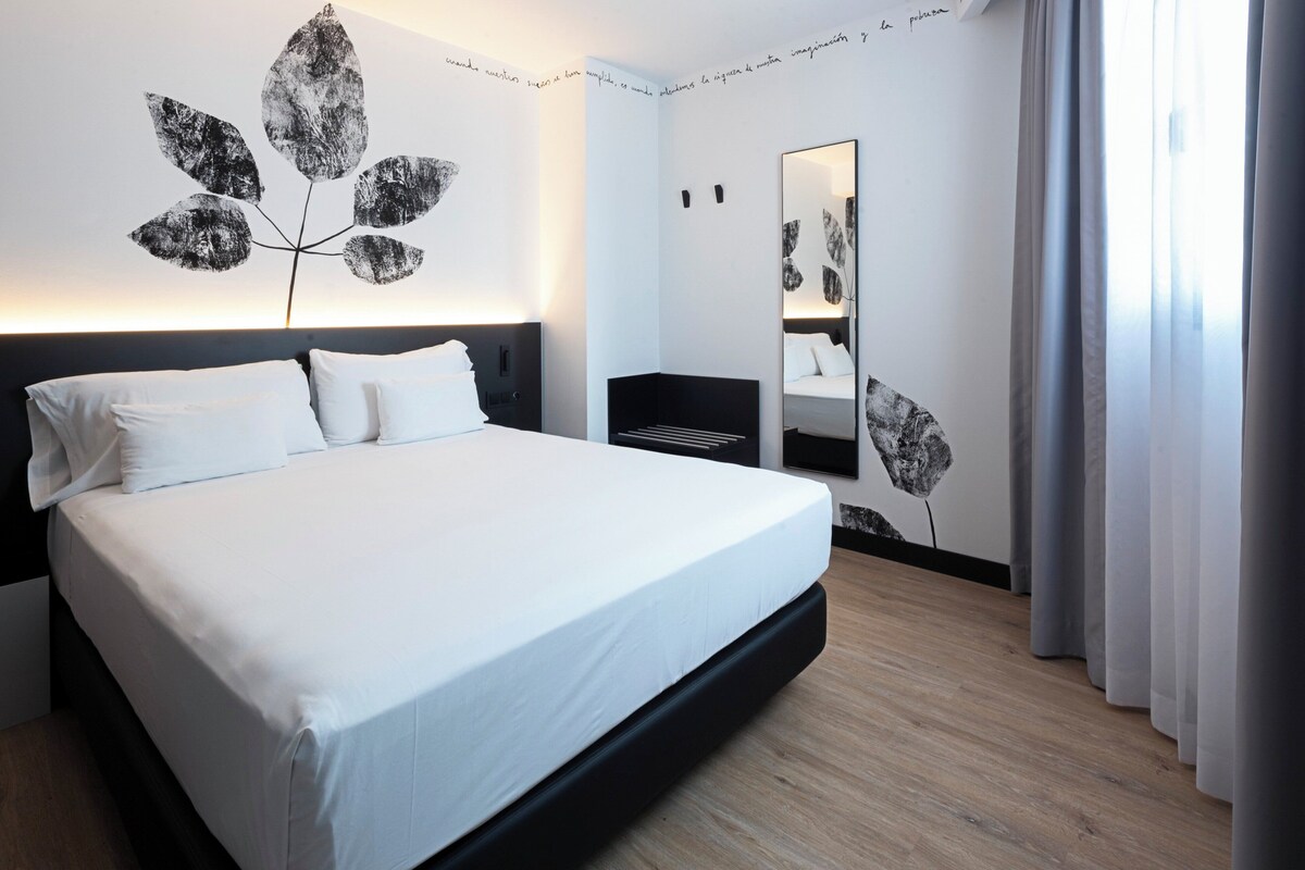A modern hotel room with a large bed, a bedside table, a mirror, and a wall art piece.