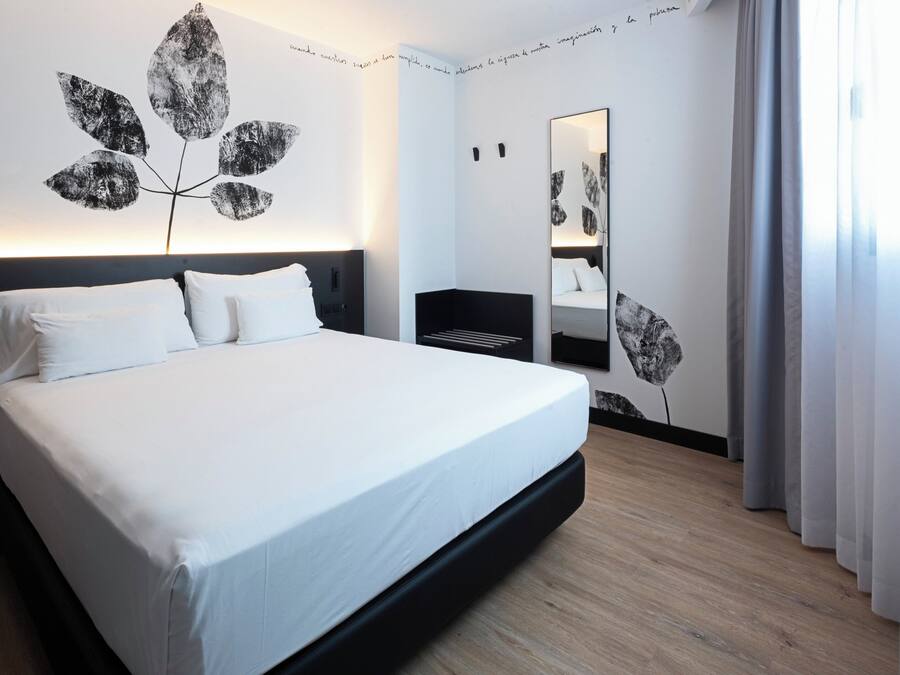 A modern hotel room with a large bed, a bedside table, a mirror, and a wall art piece.