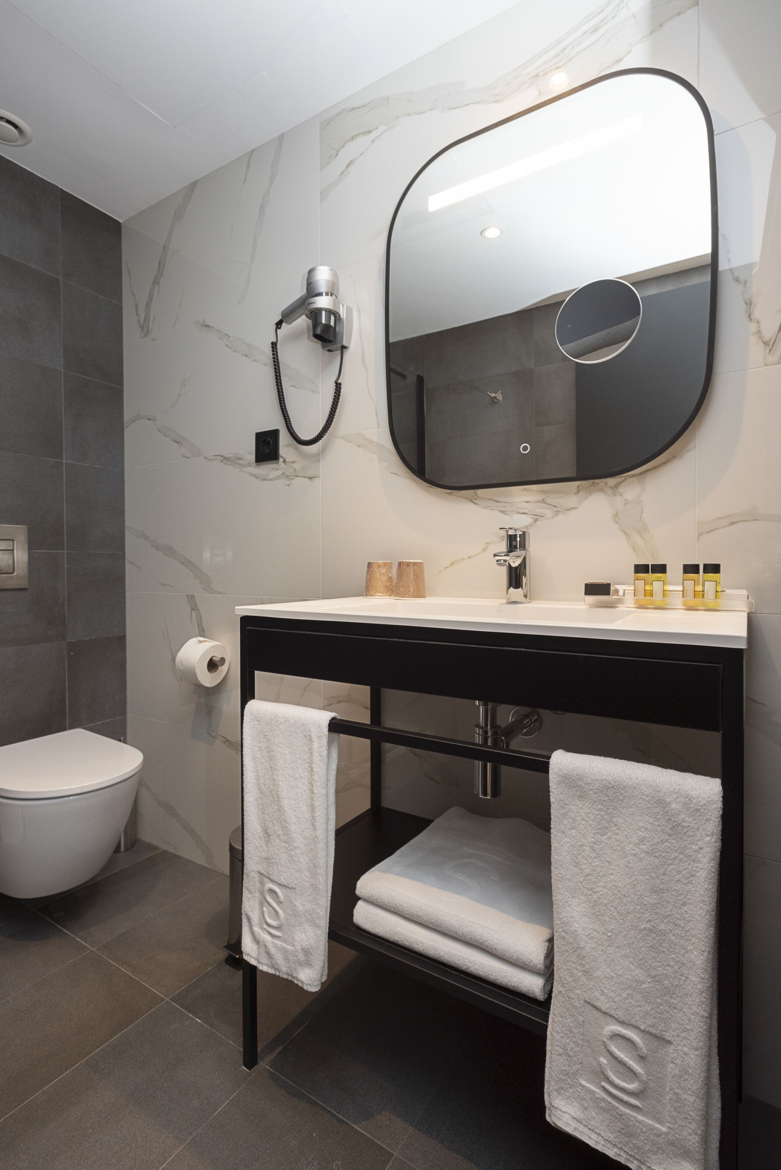 Un baño moderno con pared de mármol, tocador negro, un espejo grande y un inodoro.