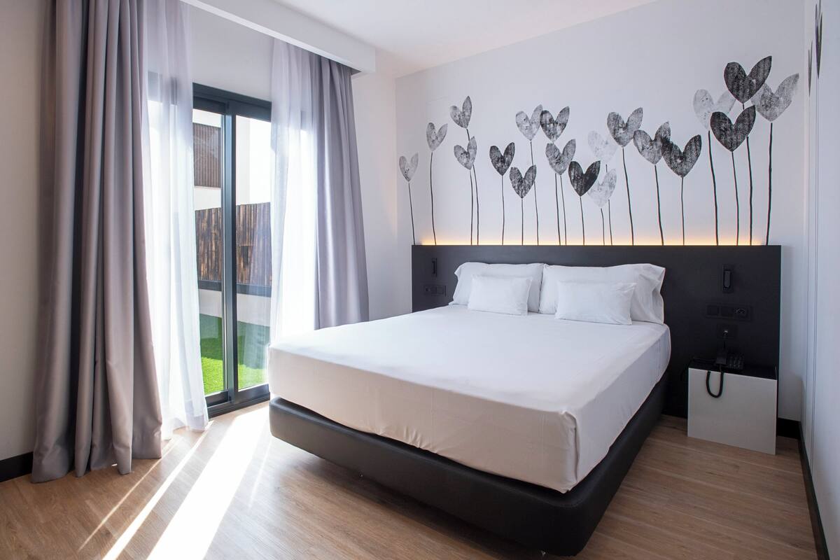 Habitación de hotel moderna con una cama grande, una mesita de noche, teléfono en la pared y vistas a un balcón.