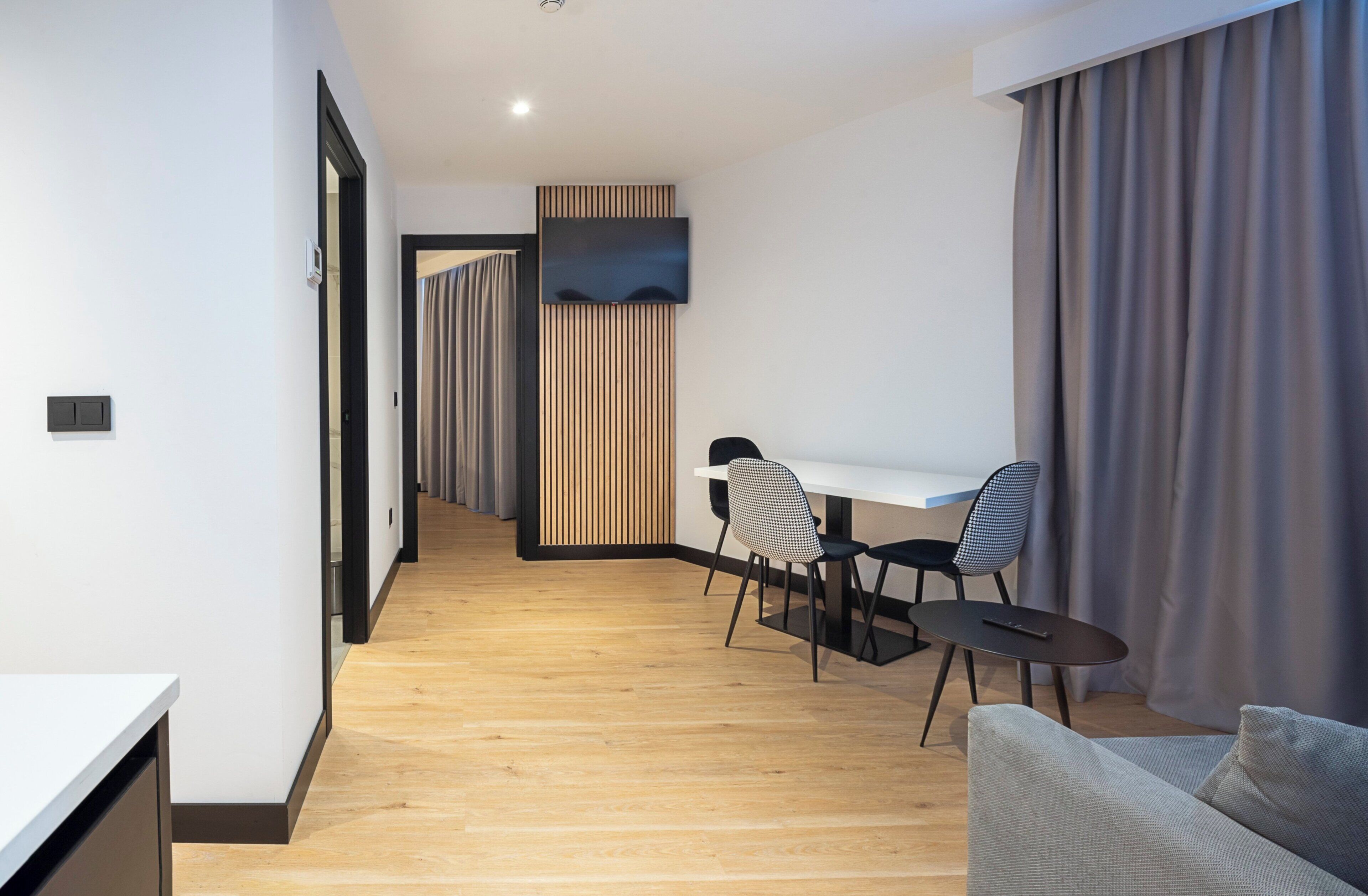 A modern hotel room with a wooden floor, a white desk, two chairs, a sofa, and a TV mounted on the wall.