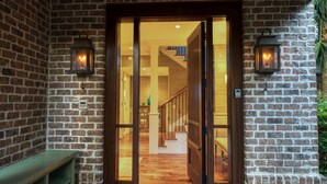 Une façade en briques avec une porte en bois donnant sur un intérieur comprenant un escalier et un banc.