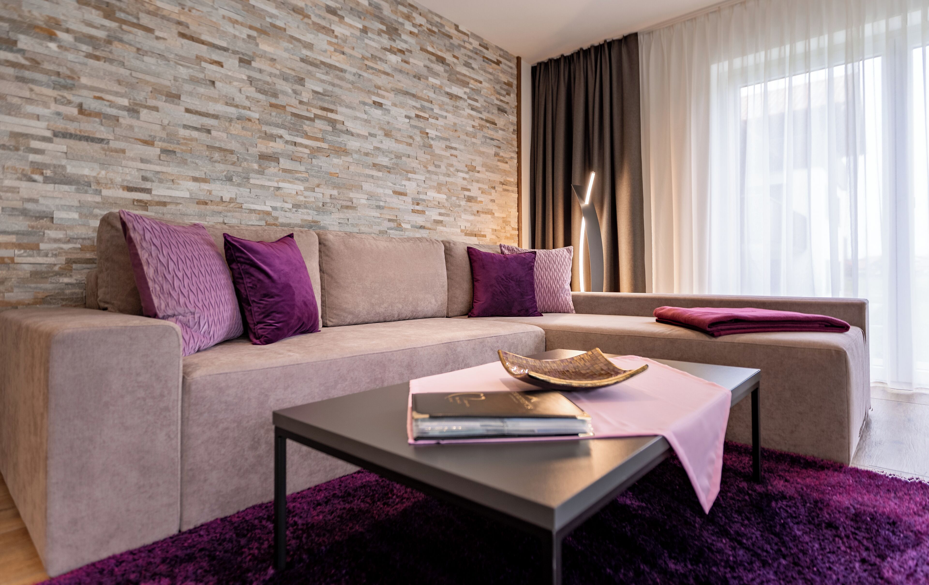 A modern living room with a stone wall, a beige sofa, purple cushions, a coffee table with a book and a woven basket, and a view of the outdoors through sheer curtains.