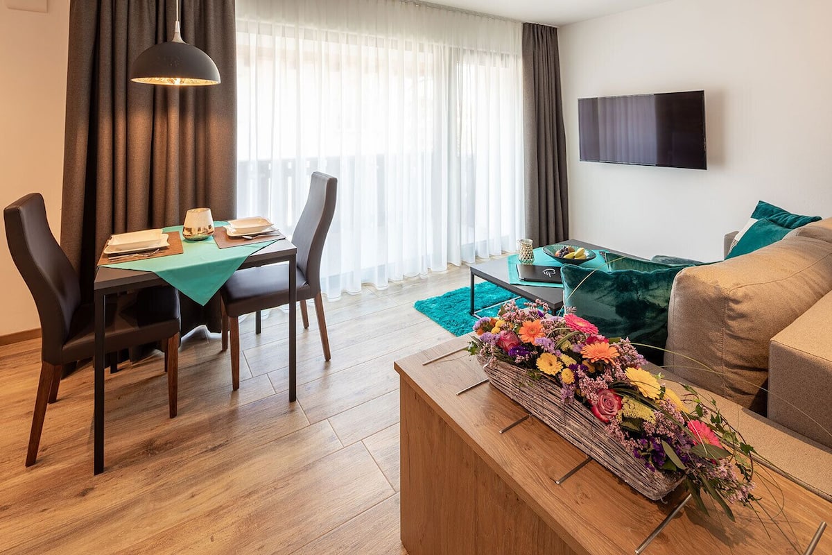 A modern living room with a dining area, a flat-screen TV mounted on the wall, and a floral arrangement on a wooden coffee table.