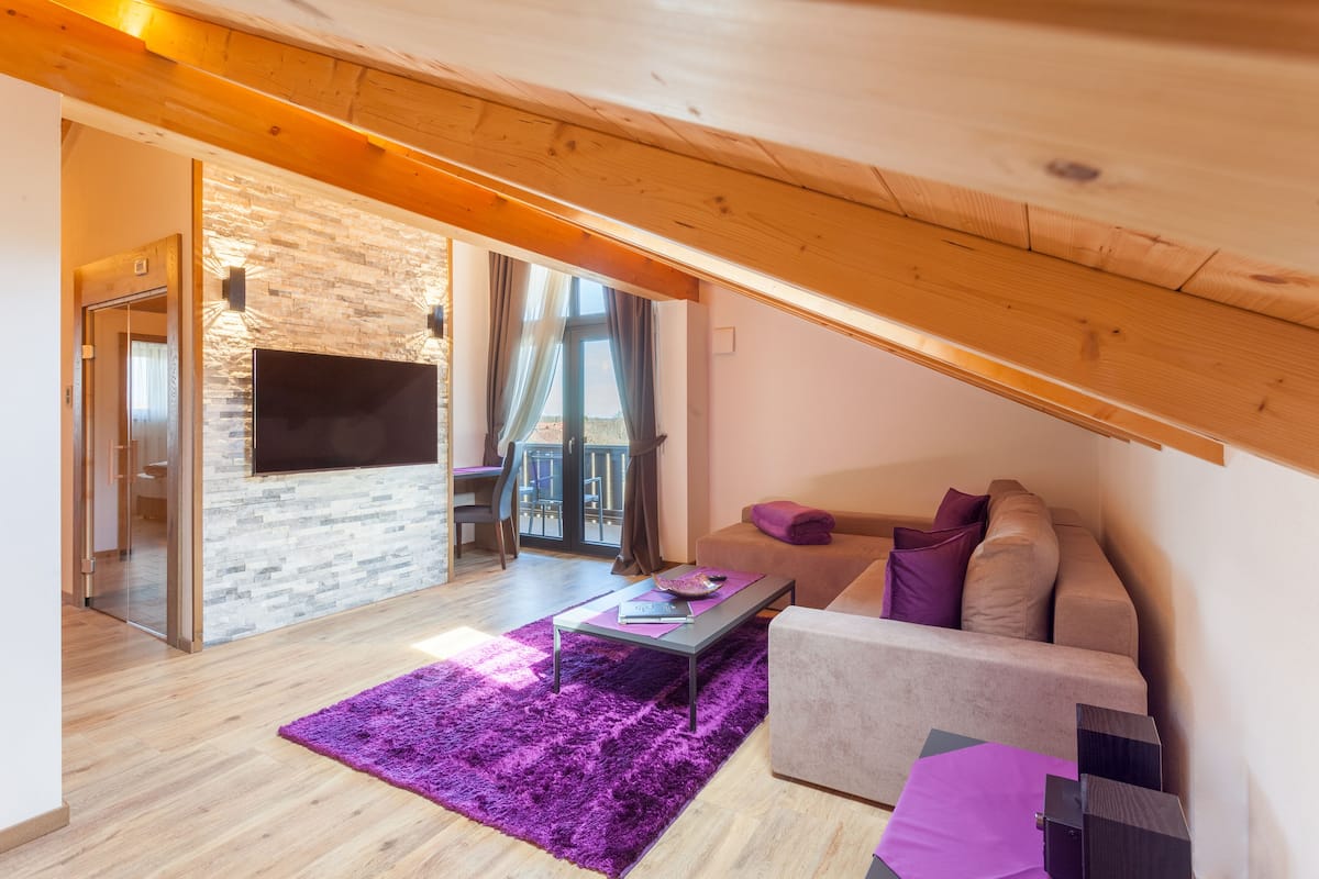 A modern living room with a stone wall, wooden ceiling beams, a flat-screen TV, a sofa with cushions, and a purple rug.