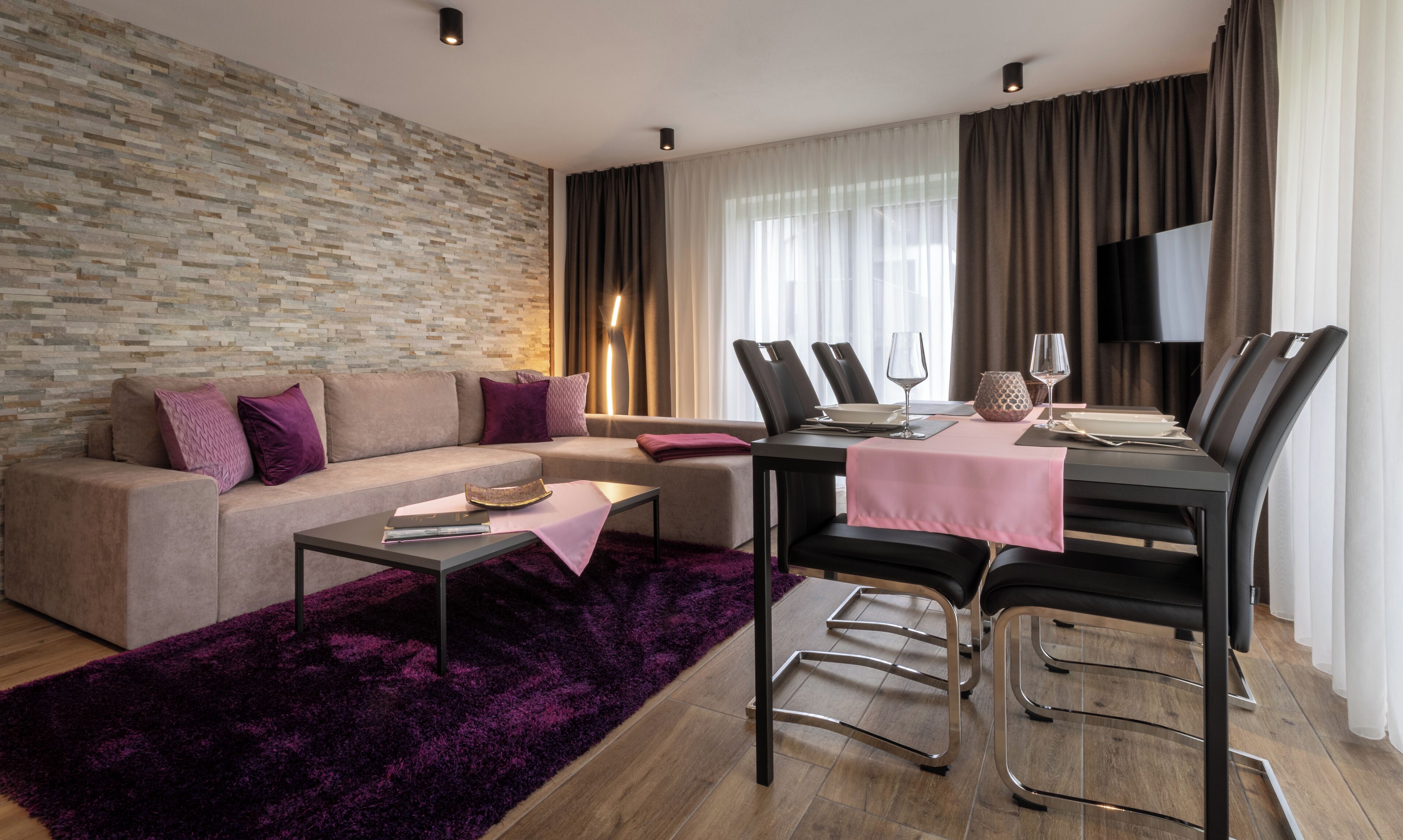 A modern living room with a stone wall, a sofa, a dining table with chairs, a coffee table, and a TV.