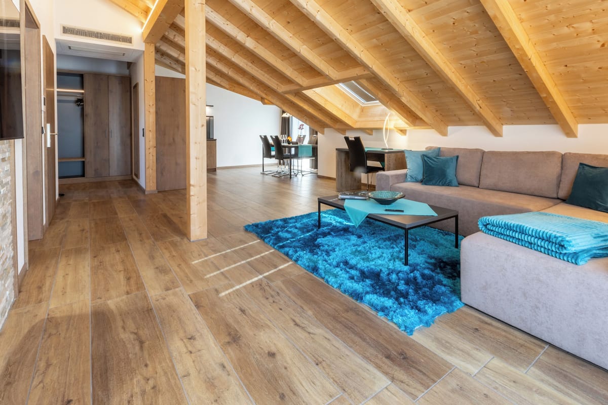 A spacious loft with a high ceiling, wooden beams, a large sofa, a dining area, and a kitchen in the background.