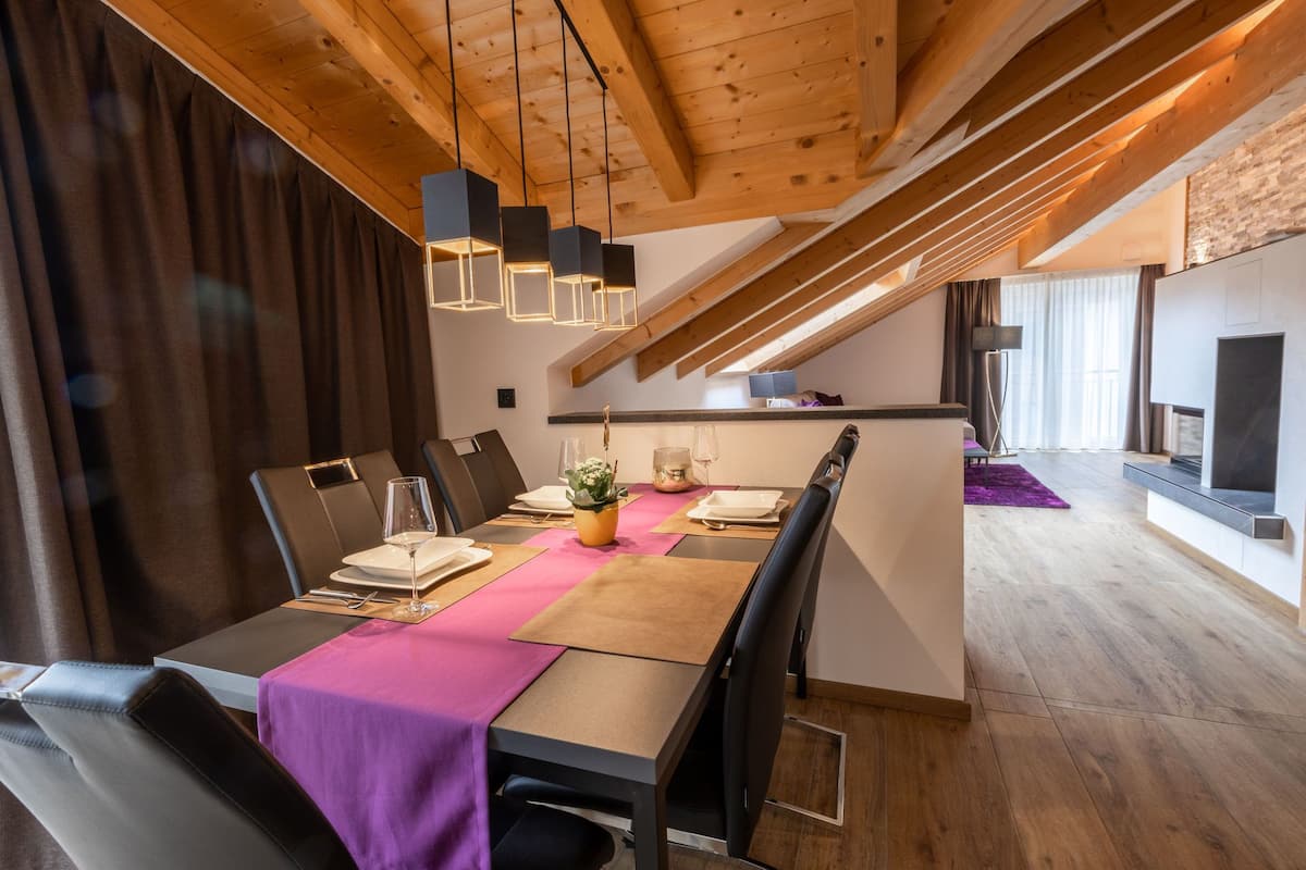 A dining area with a long table, chairs, and a purple table runner. There are hanging pendant lights and a wooden ceiling.