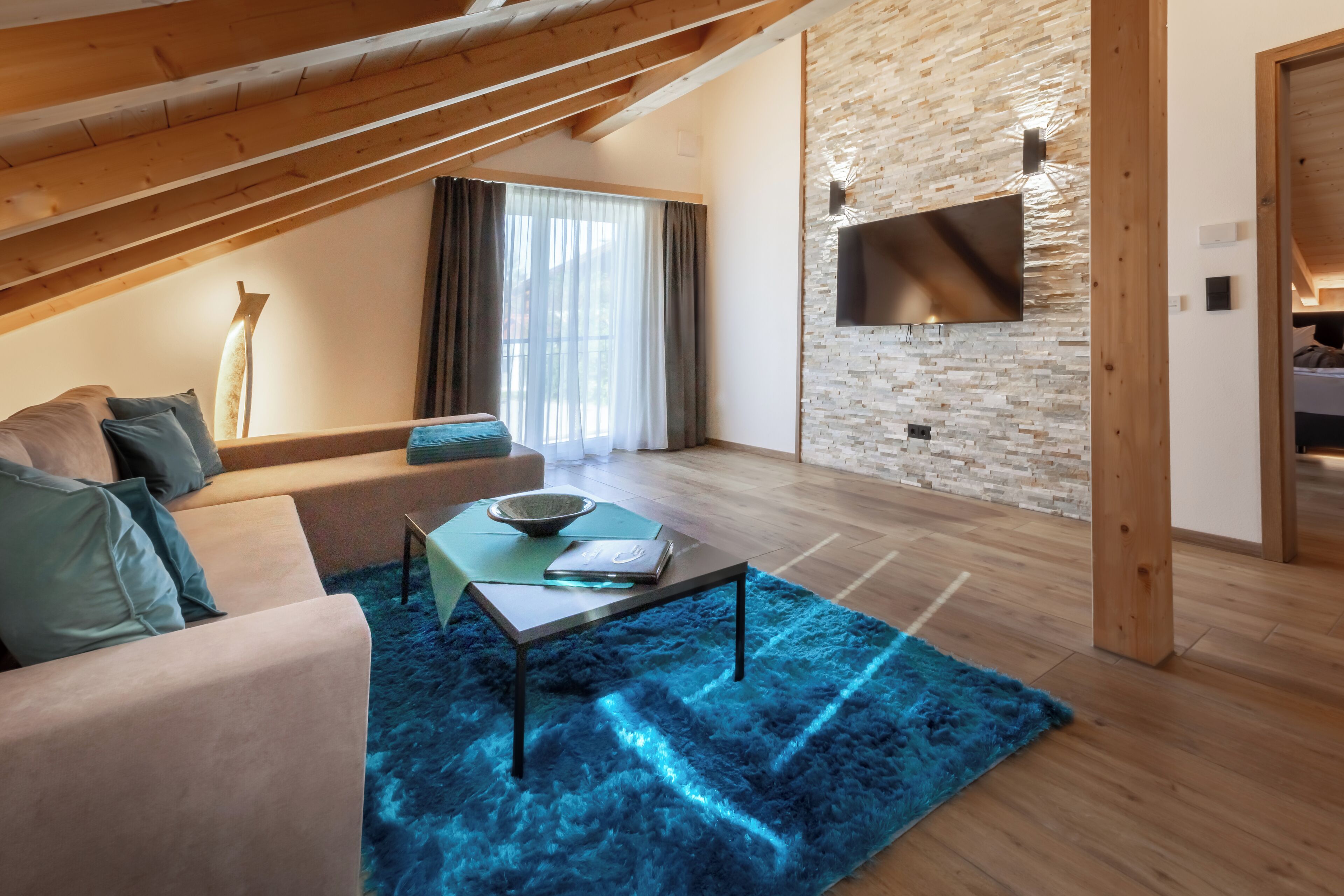 A modern living room with a sloped ceiling, a flat-screen TV mounted on a brick wall, a sofa with cushions, a coffee table, and a blue rug.