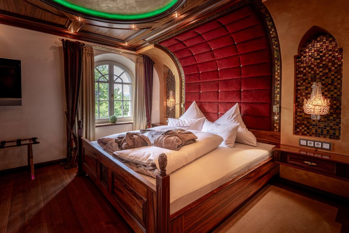 A luxurious bedroom with a large bed, wooden headboard, and a view of greenery outside.