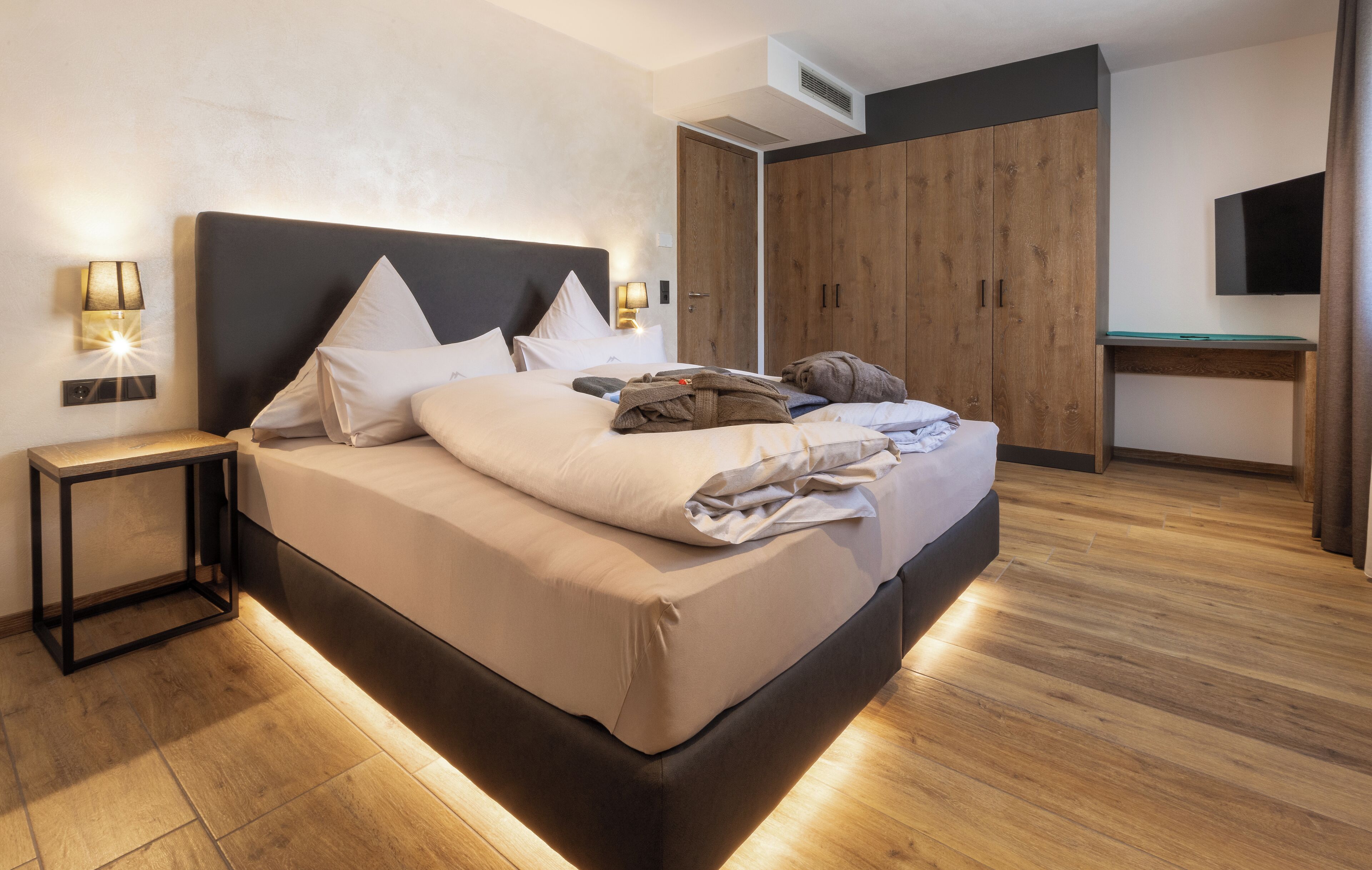 A modern bedroom with a large bed, bedside lamps, a wooden wardrobe, and a flat-screen TV.
