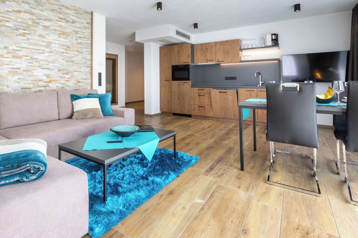 A modern living room with a sofa, coffee table, and a blue rug. There is a kitchen area with wooden cabinets and a dining table.