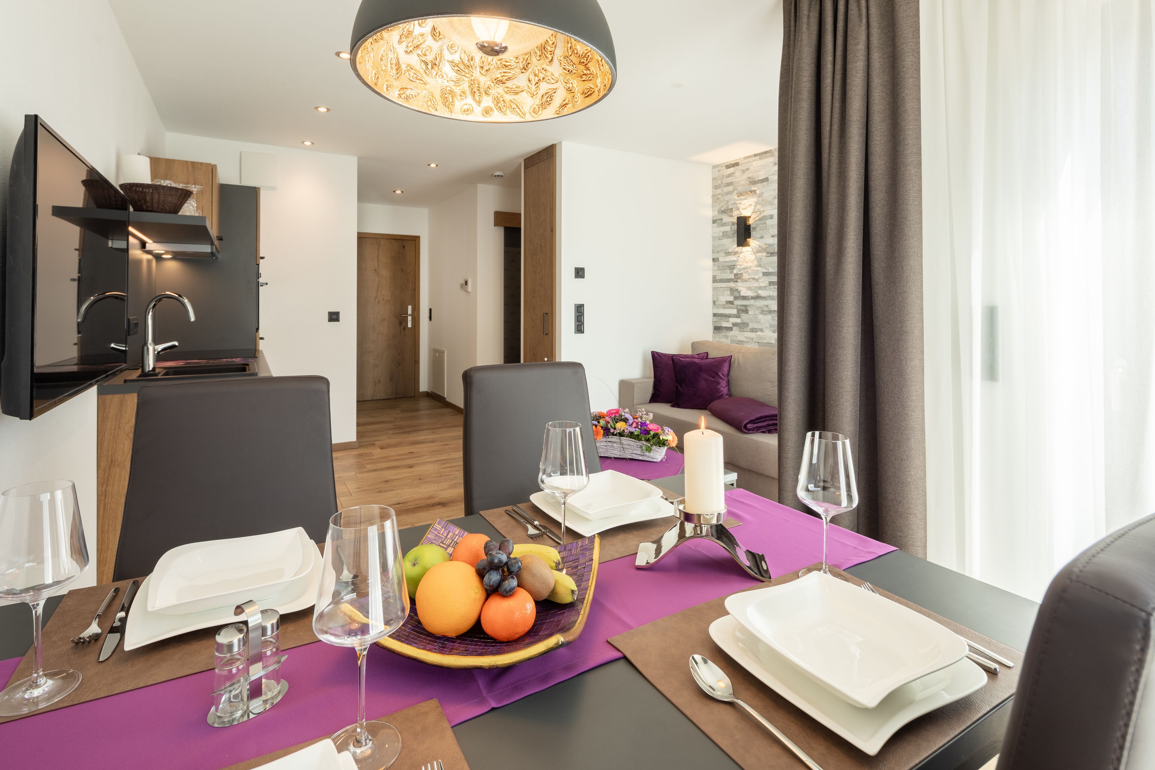 A modern dining area with a table set for two, a fruit bowl, candles, and a view of the living room.