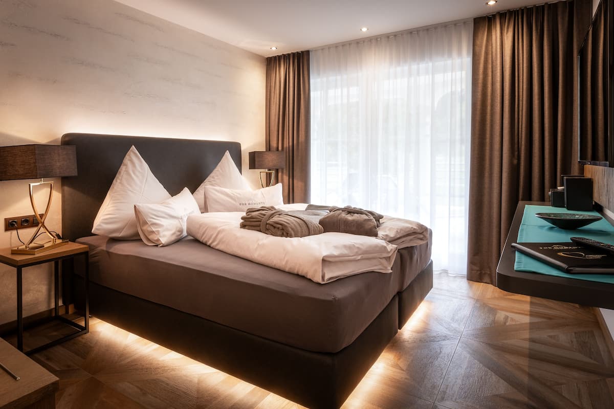 A modern hotel room with a large bed, two bedside tables with lamps, a flat-screen TV, and a view of the outdoors through sheer curtains.