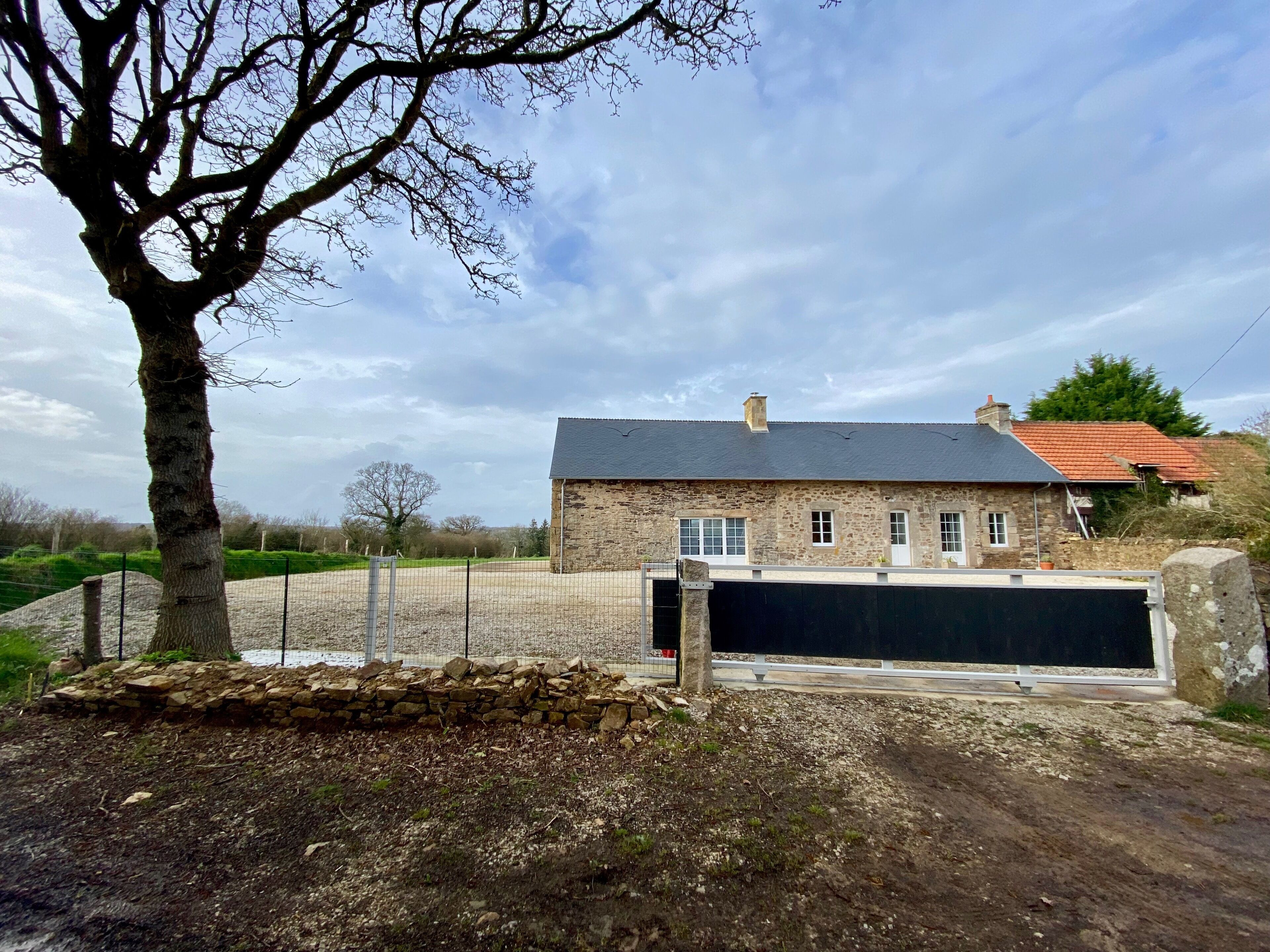 A stone building with a red-tiled roof, a fenced driveway, and a large tree in the foreground.