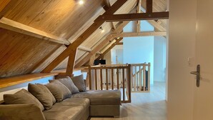 An attic bedroom with a wooden ceiling, a wooden railing, and a beige sofa with cushions. - Gîte 12/14 pers. in the countryside 10km from Barfleur and Saint-Vaast-la-Hougue (Teurthéville-Bocage)