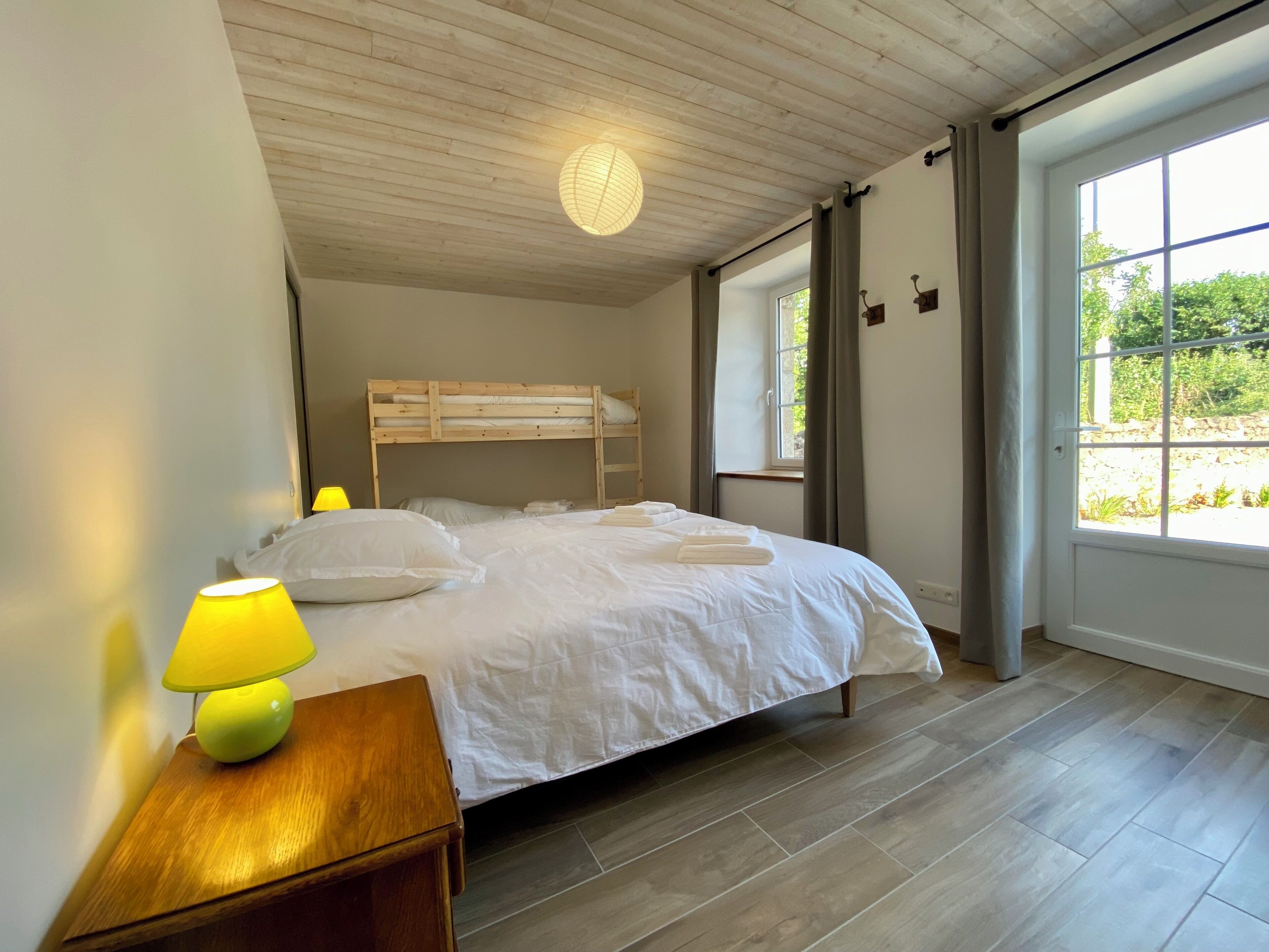 A bedroom with a bunk bed, a bedside table with a lamp, a window with curtains, and a view of greenery outside.